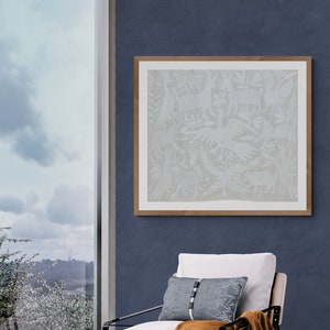 May include: A framed square wall art piece featuring a light gray intricate animal and foliage pattern, displayed above a chair with a white cushion, a blue rectangular pillow with a central vertical strap, and a draped brown throw blanket.