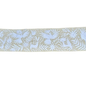 Puede incluir: Un camino de mesa rectangular de color beige con bordados blancos de aves, ciervos y diversos follajes tropicales. El intrincado trabajo de aguja cubre toda la superficie, mostrando un patrón repetitivo de fauna y plantas en tonos claros.