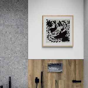 May include: A square framed wall art piece featuring a black and white illustration of a rabbit, a bird, and a butterfly surrounded by floral and leaf motifs. The artwork is enclosed in a light-colored wooden frame and mounted on a white wall.