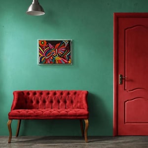 Puede incluir: Un sofá capitoné rojo con patas de madera curvadas colocado frente a una pared verde. Sobre el sofá cuelga una obra de arte rectangular enmarcada que presenta un diseño bordado vibrante y multicolor de aves y flores sobre fondo negro.