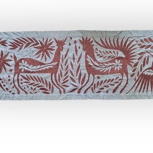 May include: A rectangular light gray fabric runner featuring an intricate embroidered pattern in a deep reddish-brown thread. The design displays two central deer figures facing each other, flanked by large birds with spread wings and floral motifs.