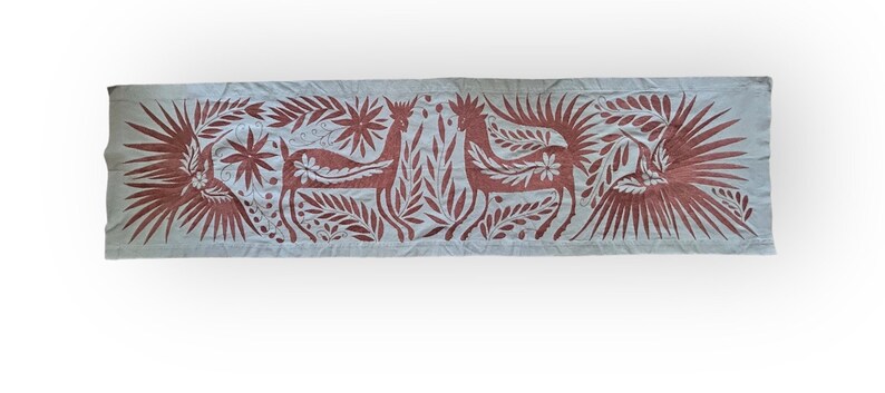 May include: A rectangular light gray fabric runner featuring an intricate embroidered pattern in a deep reddish-brown thread. The design displays two central deer figures facing each other, flanked by large birds with spread wings and floral motifs.