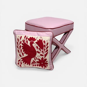 Puede incluir: Un taburete rosa claro con base en forma de X y un cojín cuadrado a juego que presenta un panel central color crema con bordado rojo oscuro que representa un ave rodeada de motivos florales y de hojas, con ribetes rojo oscuro en ambos.