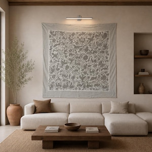 May include: A large square wall tapestry featuring a dense, intricate pattern of gray birds, animals, and foliage on a light beige fabric background. The tapestry is mounted on a wall above a light-colored sectional sofa and a wooden coffee table.