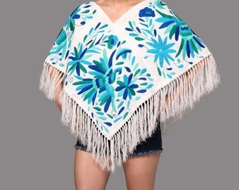 May include: A white poncho featuring a V-neckline and long fringe trim along the bottom edge. The garment is decorated with intricate floral and bird embroidery in various shades of blue, teal, and green, covering the front surface area.
