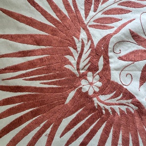 May include: A light gray fabric textile featuring intricate embroidery in a deep reddish-brown thread. The design depicts a large bird with expansive, detailed wings and a small five-petaled flower centered on its body, surrounded by leaf motifs.