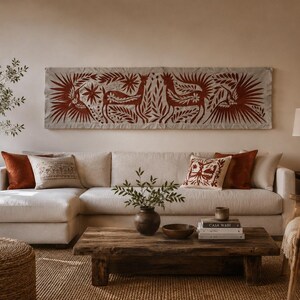 May include: A long rectangular wall hanging featuring a rust-colored folk art design of deer and birds on a light fabric background, positioned above a large off-white sectional sofa with several solid and patterned throw pillows in neutral tones.