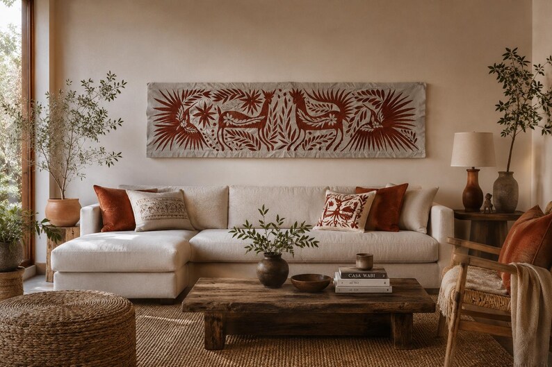May include: A long rectangular wall hanging featuring a rust-colored folk art design of deer and birds on a light fabric background, positioned above a large off-white sectional sofa with several solid and patterned throw pillows in neutral tones.