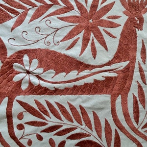 May include: A light gray fabric featuring dense, dark brown embroidery of a stylized animal, possibly a deer, surrounded by intricate floral and leaf patterns. A small, white, six-petaled flower is stitched onto the body of the animal.