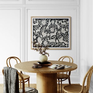 May include: A framed rectangular wall art piece featuring a black background with white folk-art style illustrations of birds, rabbits, and floral patterns. The artwork is displayed in a light wood frame on a white wall above a round wooden dining table.