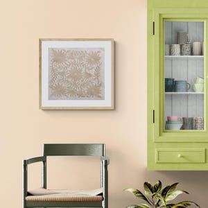 May include: A square framed wall art piece featuring a light beige floral pattern on a white background, displayed next to a lime green cabinet containing various ceramic mugs and a small green plant in the foreground.