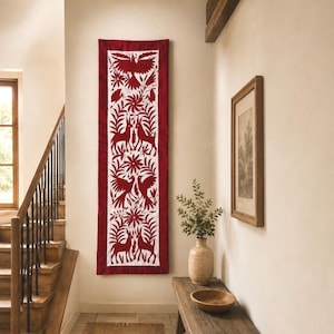 Puede incluir: Un tapiz vertical largo que presenta un panel de tela blanca con un intrincado bordado rojo. El diseño muestra patrones simétricos de aves, ciervos y motivos florales, enmarcados por un borde de tela roja sólida en los cuatro lados.
