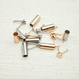 Op de afbeelding: Een verzameling metalen sieradenonderdelen, waaronder goud- en zilverkleurige cilindrische koordeinden en kleine metalen sluitingen met platte ronde voetjes en dunne draadlussen, verspreid over een gestructureerde, gebroken witte stoffen ondergrond.