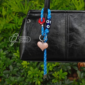 Puede incluir: Un charm de cordón azul y blanco con un colgante en forma de corazón rosa, una flor roja y cuentas azules con forma de ojo. El charm está sujeto a un bolso negro. El logotipo de la marca "Charm Polly" es visible.