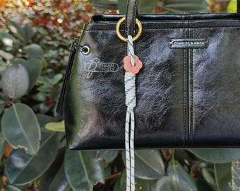 May include: Black handbag with a glossy finish, featuring a decorative charm with a pink flower and white and gray cord. The bag has a top handle and the brand name "CHARLES & KEITH" is visible.