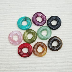 Puede incluir: Una colección de nueve cuentas en forma de rosquilla en varios colores, incluyendo turquesa, morado, negro, rosa, verde, rojo y marrón. Las cuentas tienen un acabado brillante y están dispuestas sobre una superficie blanca.