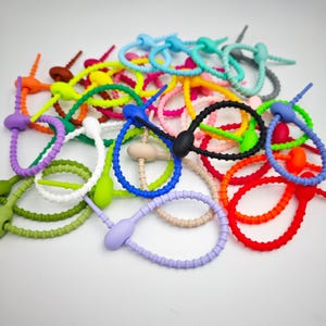 Puede incluir: Una colección de bridas de silicona de colores en varios colores, incluyendo rojo, naranja, verde, azul, morado y negro. Cada brida tiene un lazo y una pestaña de seguridad. Estas bridas reutilizables son ideales para organizar cables.