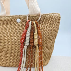 Peut inclure: Un sac fourre-tout en paille tressée avec une anse crème et un anneau décoratif retenant plusieurs pompons en corde colorés. Les pompons sont dans les tons de rouge, crème, marron et jaune, ajoutant une touche de couleur au sac neutre.
