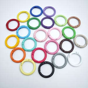 Könnte beinhalten: Eine Sammlung farbenfroher, runder Metall-Federringverschlüsse in verschiedenen Farben, darunter Rot, Gelb, Blau, Grün, Rosa und Schwarz. Diese Verschlüsse sind zum Öffnen und Schließen konzipiert und eignen sich zum Basteln oder Befestigen von Gegenständen.