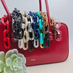 May include: A red handbag with a selection of colourful chain straps. The chains are in shades of red, black, white, silver, blue, green, brown, and clear. The handbag is rectangular and has the word "muva" printed on the front.