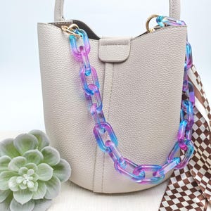 Puede incluir: Un bolso gris claro con una colorida correa de cadena en tonos azules y morados. El bolso tiene un asa pequeña y una bufanda decorativa con un estampado a cuadros marrón y blanco.