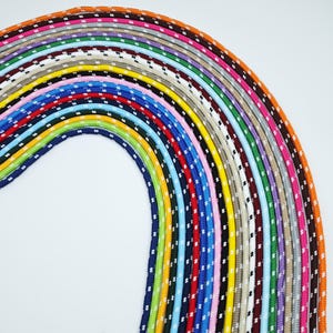 Könnte beinhalten: Eine Sammlung farbiger Kordeln, jede mit einem einzigartigen Muster aus weißen Punkten. Die Kordeln sind in einer gebogenen, regenbogenartigen Anordnung angeordnet und zeigen eine Vielzahl von Farben, darunter Blau, Grün, Rot und Orange. Die Kordeln werden wahrscheinlich zum Basteln oder für Schmuck verwendet.