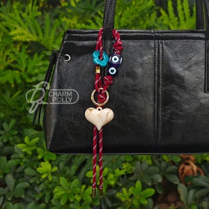 May include: A decorative bag charm with a heart-shaped pendant and a red and white cord. The charm also features turquoise and blue beads, and a gold-colored ring. The charm is attached to a black handbag.