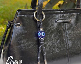 May include: A black handbag with a decorative charm. The charm features a blue glass bead with white eye details, suspended from a black and white cord. The handbag has a glossy finish and the brand name "CHARLES & KEITH" is visible.