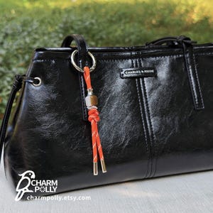 Peut inclure: Sac à main en cuir noir avec un charm en corde orange et blanc. Le charm comprend une perle métallique et des accents dorés. Le sac à main a une finition brillante et la marque "CHARLES & KEITH" est visible.