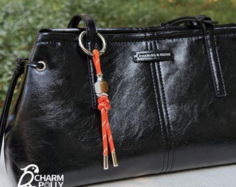 May include: Black leather handbag with a decorative orange and white rope charm. The charm features a metal bead and gold-colored accents. The handbag has a shiny finish and the brand name "CHARLES & KEITH" is visible.