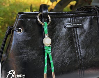 May include: A black leather handbag with a green and white charm. The charm features a white bead and green and white cord with gold-colored metal ends. The handbag has a silver ring attached to the handle.
