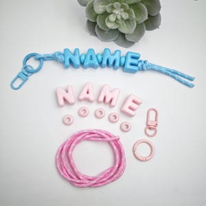 May include: A collection of personalized accessories, including a blue keychain spelling "NAME", pink letter beads, small pink rings, a pink clasp, and a pink and white rope. A succulent plant is in the background.