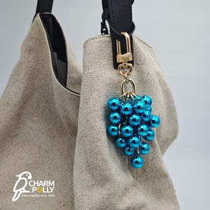 Puede incluir: Un charm metálico azul en forma de racimo de uvas cuelga de un cierre dorado sujeto a una correa negra. El charm está colgado de un bolso tote beige. El nombre de la tienda Etsy "Charm Polly" es visible en la esquina inferior izquierda.