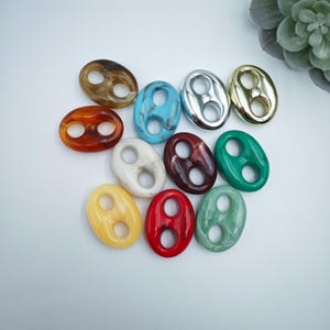 Könnte beinhalten: Eine Sammlung ovaler Knöpfe in verschiedenen Farben, darunter Braun, Türkis, Silber, Gold, Weiß, Burgunderrot, Grün, Gelb, Rot und marmoriertes Grün. Jeder Knopf hat zwei Löcher. Die Knöpfe sind auf einer weißen Oberfläche angeordnet.