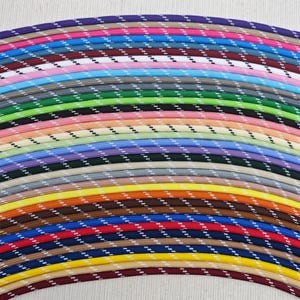 Puede incluir: Una colección de coloridos cordones trenzados en varios tonos, incluyendo morado, rosa, azul, verde y amarillo. Cada cordón presenta un patrón diagonal blanco y negro, creando una apariencia texturizada. Los cordones están dispuestos en una formación curva y en capas.