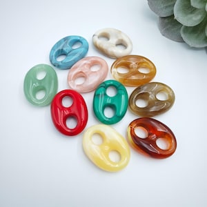 Puede incluir: Una colección de cuentas ovaladas de colores con dos agujeros, en tonos de azul, blanco, rosa, verde, rojo, amarillo y marrón. Las cuentas son lisas y brillantes, y están dispuestas sobre una superficie blanca.