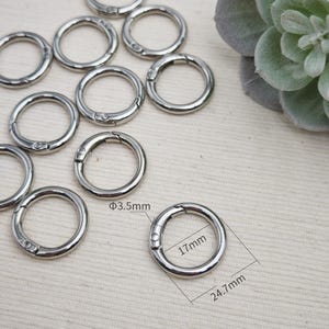Può includere: Una collezione di anelli a molla in metallo color argento. Un anello è contrassegnato con dimensioni che indicano uno spessore di 3,5 mm, un diametro interno di 17 mm e un diametro esterno di 24,7 mm. Questi connettori circolari hanno un meccanismo a molla.