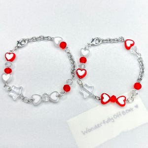 May include: Two silver chain bracelets with red heart and bead accents. One bracelet features a bow charm, while the other has star-shaped charms. The bracelets are adorned with clear, faceted beads and white heart-shaped charms.