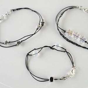 May include: Three delicate bracelets with black cords and silver-toned clasps. One bracelet features star-shaped charms, another has iridescent star-shaped beads, and the third has a single bead. The bracelets are displayed on a white surface.