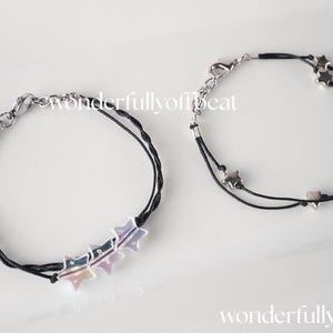May include: Two black cord bracelets with silver-toned metal hardware. One bracelet features three iridescent star charms, while the other displays two small silver star charms. Both bracelets include a lobster claw clasp and chain extender.
