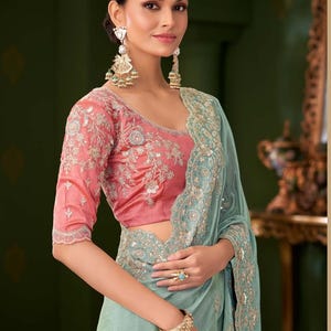 May include: A woman wearing a traditional Indian sari. The sari features a light blue drape with intricate embroidery and a pink blouse with floral embroidery. She is wearing gold earrings, a bracelet, and a ring.