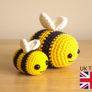 May include: Two crocheted bumblebees, one larger than the other, with black and yellow stripes and white wings. The smaller bee is in front of the larger bee.