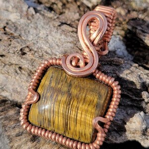 May include: A copper wire wrapped pendant featuring a square-shaped tiger's eye gemstone. The pendant has a copper wire bail with a decorative swirl design.