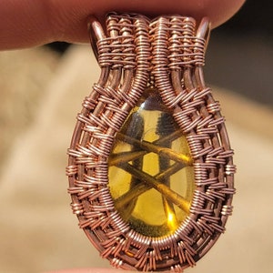 May include: A copper wire wrapped pendant with a yellow-brown gemstone in the center. The wire is woven in a basket weave pattern.