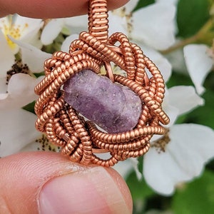May include: A handcrafted copper wire-wrapped pendant featuring a rough-cut, lavender-colored gemstone. The wire is intricately woven around the stone, creating a cage-like design. The pendant is held between fingers against a backdrop of white flowers.