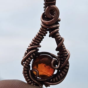 May include: A teardrop-shaped pendant crafted from coiled copper wire, featuring a central round amber-colored gemstone. The wire is intricately wrapped around the stone, forming a secure setting with a loop at the top for a chain or cord.