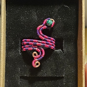 May include: A pink and purple wire-wrapped snake ring with a green stone for the eye. The ring is displayed in a black velvet box.