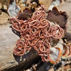 May include: A copper wire star-shaped sculpture with intricate swirls and loops. The sculpture is resting on a piece of wood with a light brown color.