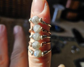 May include: Five handmade rings with silver and copper wire, each featuring a small, round, gray gemstone. The rings are stacked on a finger, showcasing the jewelry's design and craftsmanship. The gemstones have a subtle, iridescent quality.