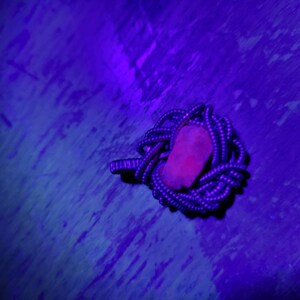 Op de afbeelding: Een kleine, rozerode steen ligt genesteld in een spoel van donkerblauw en wit gespikkeld touw. De afbeelding wordt verlicht met een paarse tint, waardoor een contrast ontstaat tussen de steen en het touw.